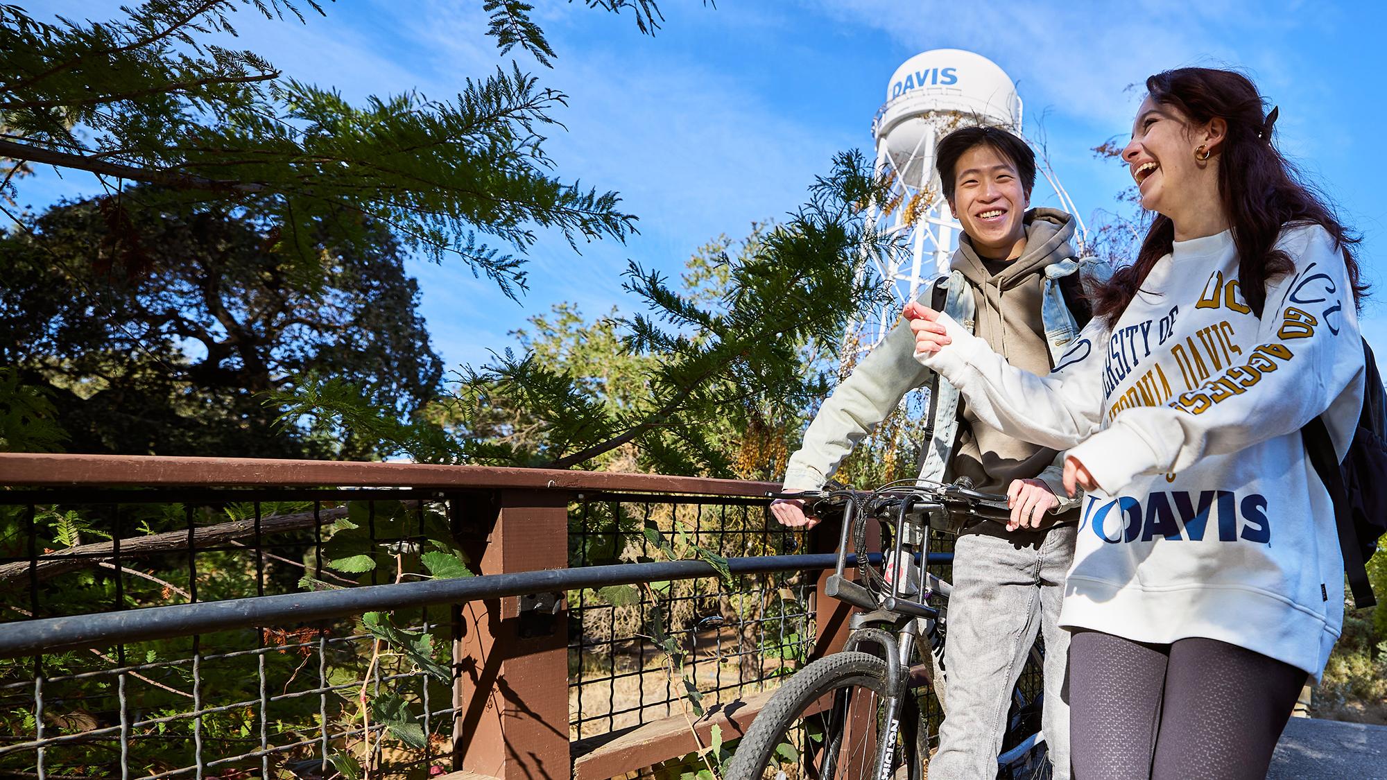 Two students smile and walk across a bridge on the UC Davis campus. One pushes a bicycle and both wear UC Davis sweatshirts. The campus water tower appears in the background.